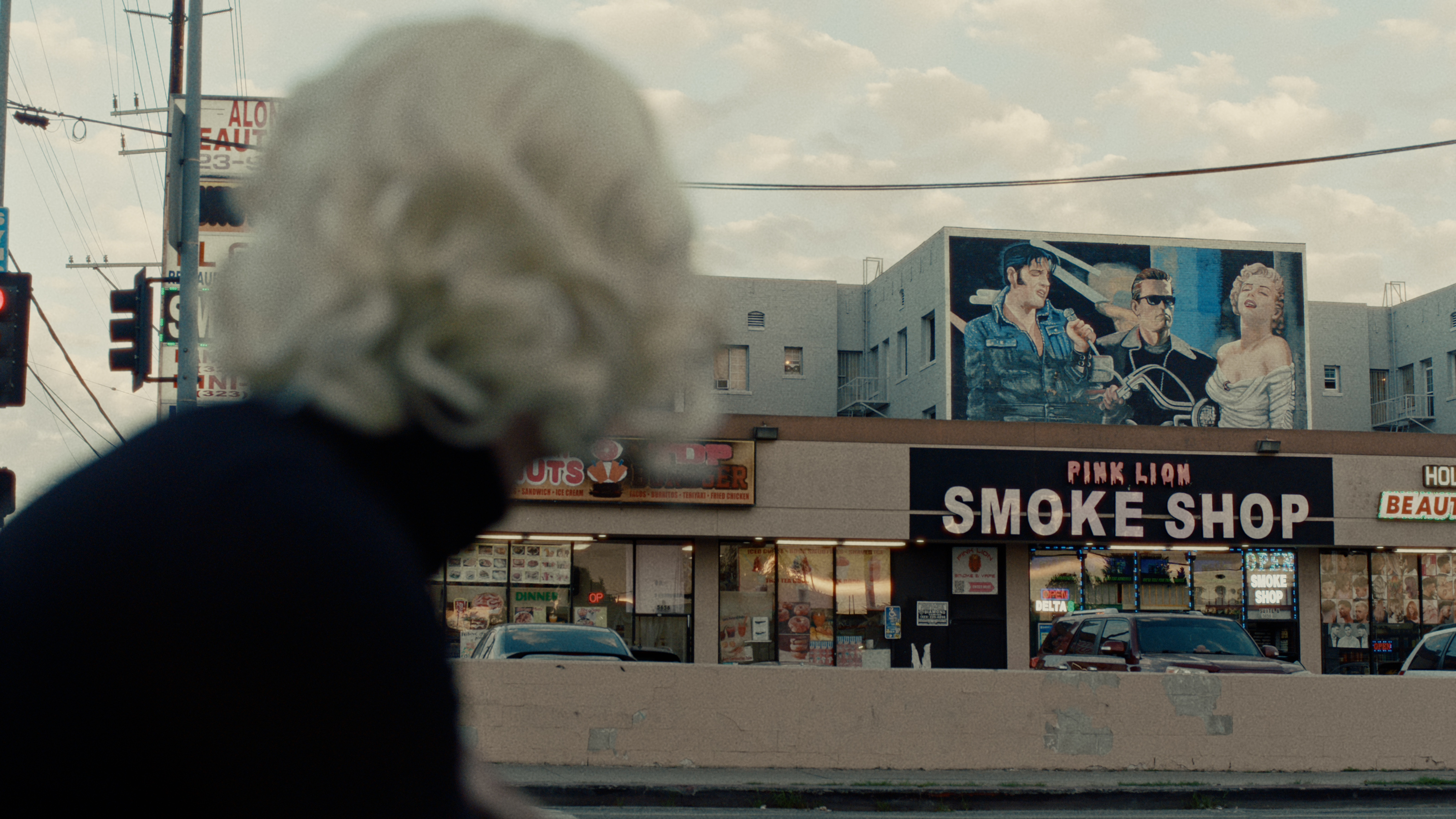 A blonde figure gazes at a mural of Elvis, James Dean, and Marilyn Monroe above a smoke shop