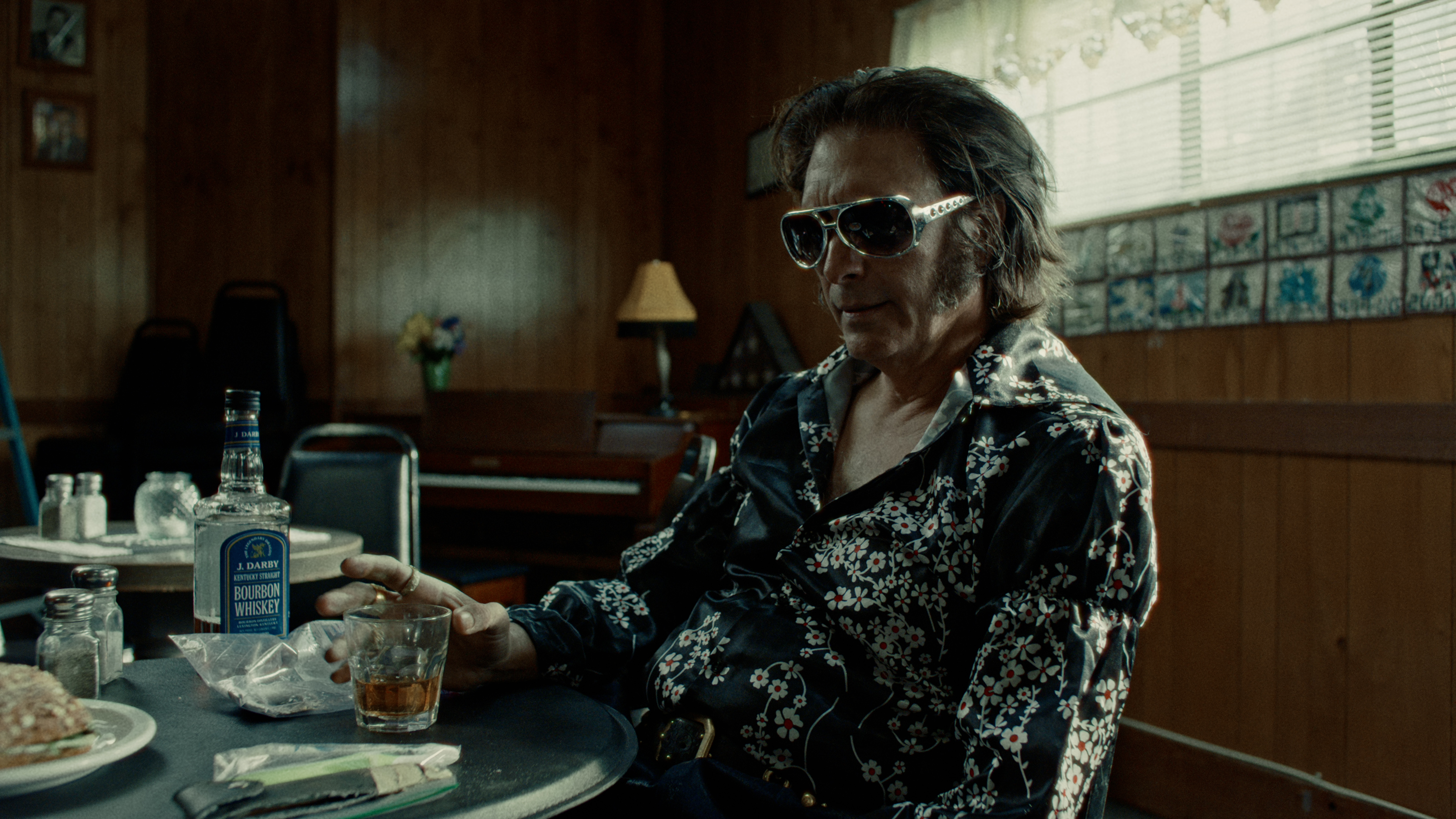 A man in rhinestone sunglasses and a floral silk shirt holds a bourbon glass